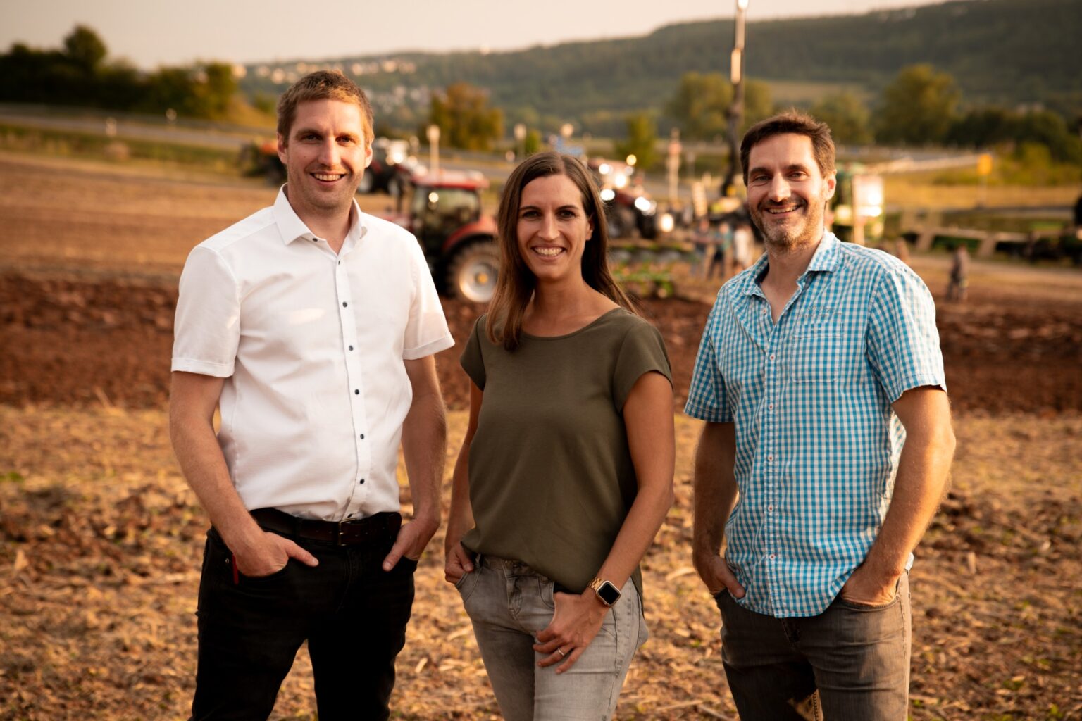 Drei Personen stehen auf einem Feld, im Hintergrund sind Traktoren und landwirtschaftliche Arbeiten sichtbar.